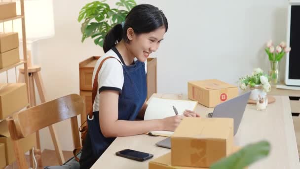 ノートやノートパソコンを使ったアジア系女性起業家の小規模スタートアップと中小企業経営者の肖像画で 顧客のために箱に詰め込む前に製品を整理するよう注文をチェックしている — ストック動画
