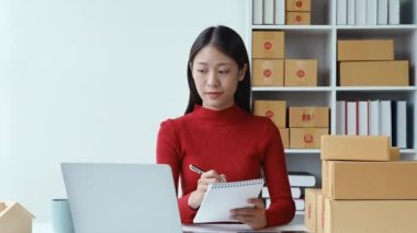 e-ticaret, küçük işletme, sme, güzel Asyalı kadın bilgisayara bakıyor ve not alıyor müşteri siparişleri alıyor ve çalışırken gülen yüzle çevrimiçi satışlardan para kazanıyor. Yüksek kalite 4k