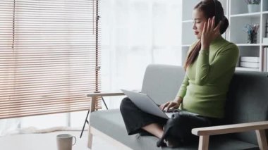 Genç Asyalı kadın kulaklık takıyor gülen yüzünü gösteriyor ve evdeki kanepede dinlenirken müzik dinlemek için bilgisayar kullanıyor. Yüksek kalite 4k görüntü