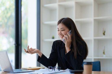 Sabahın erken saatlerinde masasında telefonunu, bilgisayarını ve mali belgelerini kullanırken gülümseyen bir Asyalı kadının portresi..