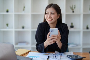 Sabahın erken saatlerinde masasında telefonunu, bilgisayarını ve mali belgelerini kullanırken gülümseyen bir Asyalı kadının portresi..