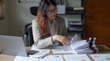 Asyalı genç bir iş kadını ya da muhasebeci. Bütçede çalışırken stresli yüzünü gösteriyor. Evrak işlerini yapıyor, bilgisayarını ahşap masaya koyuyor. Yüksek kalite 4k görüntü