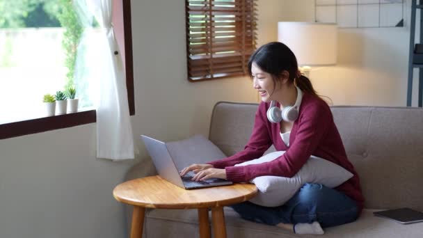 年轻的亚洲女人坐在家里的沙发上使用笔记本电脑 高质量的4K镜头 — 图库视频影像