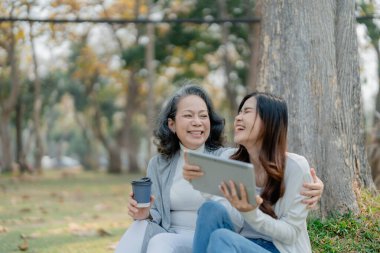 Yaşlanan bir annesi olan yetişkin bir kız çocuğu aşkı ifade ediyor ve parktaki ağacın altındaki tablet bilgisayara bakıyor.