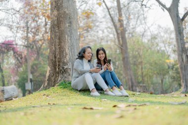 Yaşlanan bir annesi olan yetişkin bir kız. Sevgisini ifade ediyor ve parktaki ağacın altında akıllı telefonlara bakıyor.