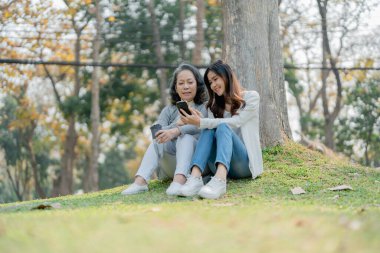 Yaşlanan bir annesi olan yetişkin bir kız. Sevgisini ifade ediyor ve parktaki ağacın altında akıllı telefonlara bakıyor.