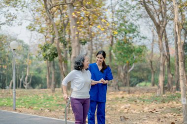 Kadın fizyoterapist yaşlı bir kadını kucağında tutuyor ve bacak kaslarını güçlendirmek için parkta yürüyor.