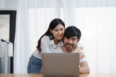 Mutlu yüzler gösteren genç evli bir çift ve evde dinlenirken ev gelirlerini ve giderlerini hesaplamak için bilgisayar ve defter kullanıyorlar.