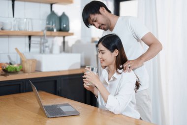 Mutlu yüzler gösteren genç evli bir çift ve evde dinlenirken ev gelirlerini ve giderlerini hesaplamak için bilgisayar ve defter kullanıyorlar.