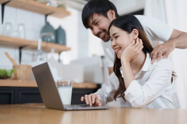 Mutlu yüzler gösteren genç evli bir çift ve evde dinlenirken ev gelirlerini ve giderlerini hesaplamak için bilgisayar ve defter kullanıyorlar.