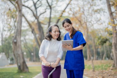 Hasta bakımı, sopayla ilgilenen genç Asyalı kadın hemşire, parkta yürürken dijital tablet kullanarak bacak ve diz egzersizleri yapıyor.