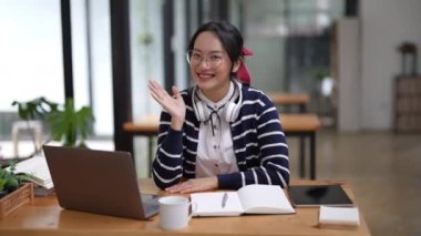 İnternette çalışmak için bilgisayar, not defteri ve tablet kullanırken gülümseyen Asyalı genç kadın el sallıyor. Yüksek kalite 4k görüntü