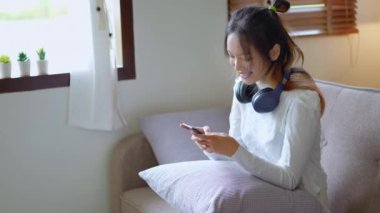 Kulaklığı kulaklıkla cep telefonu kullanan genç Asyalı kadın koltukta dinleniyor. Yüksek kalite 4k görüntü