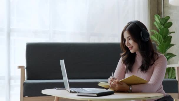 自宅のテーブルの上でコンピュータ ノートパソコン ノートパソコンを使ってヘッドフォンをしながらオンラインで勉強する魅力的な若いアジアの女性学生 高品質4K映像 — ストック動画