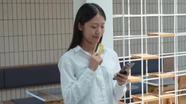Genç ve güzel Asyalı bir kadın akıllı telefon ve kredi kartını kullanarak mallara veya internet faturalarına ödeme yapıyor. Çevrimiçi ödeme ve alışveriş. Yüksek kalite 4k görüntü