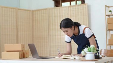 Küçük bir girişimin portresi ve KME sahibi, Asyalı bir bayan girişimci not defterleri ve dizüstü bilgisayar kullanarak müşteriler için iç kutulara koymadan önce ürünleri organize etme talimatlarını kontrol ediyor. Yüksek
