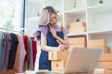 Bağımsız Asyalı kadın çevrimiçi satıcının küçük işletme girişimcisi olarak bir müşteriyle telefonda konuşuyor ve müşteriye teslimat için ürünleri paketliyor. ve KME dağıtım kavramı.