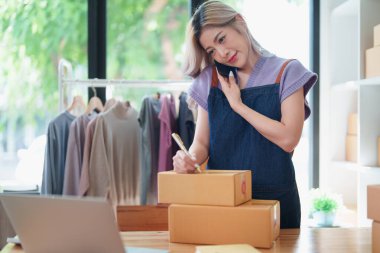 Bağımsız Asyalı kadın çevrimiçi satıcının küçük işletme girişimcisi olarak bir müşteriyle telefonda konuşuyor ve müşteriye teslimat için ürünleri paketliyor. ve KME dağıtım kavramı.