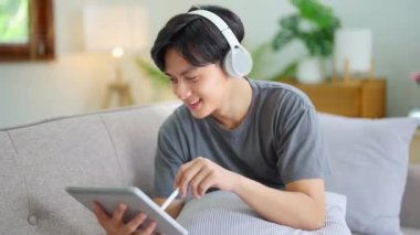 Kulaklık takan yakışıklı Asyalı adam müzik dinliyor ve tatil evinde kanepede oturup tablet kullanıyor. Yüksek kalite 4k görüntü