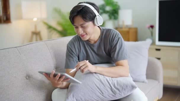 ヘッドフォンをして音楽を聴いたり 休日の家のソファに座ってタブレットを使用してハンサムなアジアの男 高品質のフルHd映像 — ストック動画