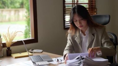 Asyalı genç bir iş kadını ya da muhasebeci. Bütçede çalışırken stresli yüzünü gösteriyor. Evrak işlerini yapıyor, bilgisayarını ahşap masaya koyuyor. Yüksek kalite 4k görüntü