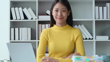 Asyalı genç bir kadın bilgisayar ve finansal dökümanlar kullanıyor, ofiste muhasebe yapıyor, kolları bağlı oturuyor, dik bakıyor, gülümseyen yüzünü gösteriyor. Yüksek kalite 4k görüntü