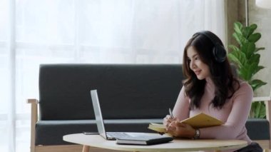 Evde bilgisayar, dizüstü bilgisayar ve dizüstü bilgisayar kullanarak kulaklık takan Asyalı çekici genç kız öğrenci. Yüksek kalite 4k görüntü