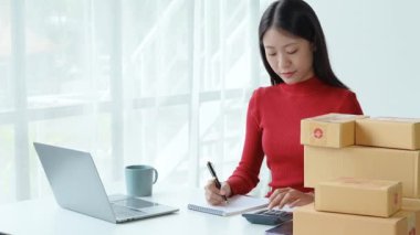 E-ticaret, küçük işletme, sme, nota bakan güzel Asyalı kadın ve hesap makinesi kullanarak müşteri siparişlerini hesaplıyor ve çalışırken gülen yüzle çevrimiçi satışlardan para kazanıyor.