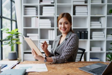 Portrait of a young Asian Woman showing a smiling face as she uses his notebook, computer and financial documents on her desk in the early morning hours.