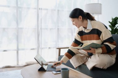 Bir kadın, bilgisayarını ve not defterini kullanarak aynı anda bir koltuğa oturur ve evindeki konfordan dijital platformlar aracılığıyla öğrenmesini sağlar..