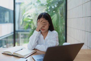 Modern ofis ortamındaki stresli genç kadın, tek eliyle yüzünü örtüyor ve bir dizüstü bilgisayar ve defterle çevrili, işin zorluklarını ve teslim tarihlerini yansıtıyor..