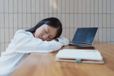 Genç bir kadın, masa başında başını kollarına dayamış, bunalmış hissediyor. Yanındaki dizüstü bilgisayar, günlük hayatta dinlenme ve denge ihtiyacını vurgulayan modern bir çalışma alanını yansıtıyor..