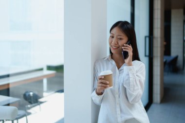 Genç bir kadın, modern, aydınlık bir ofis odasında akıllı bir telefonla konuşurken kahveden zevk alıyor..