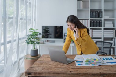 Modern ofis alanında çalışan şık sarı ceketli genç iş kadını akıllı telefon ve dizüstü bilgisayar kullanıyor..