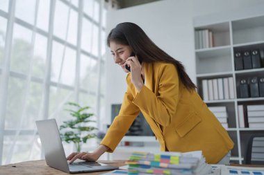 Genç iş kadını, modern ofis ortamında dizüstü bilgisayarla çalışırken doğal ışık alan bir telefon görüşmesi yaptı..