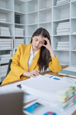 Sarı ceketli genç bir kadın iş yerinde stresli görünüyor. Evrak işleri ve modern ofis ortamlarıyla çevrili..