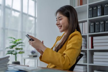 Modern ofis ortamında doğal ışık alan akıllı telefon kullanırken gülümseyen genç profesyonel kadın..