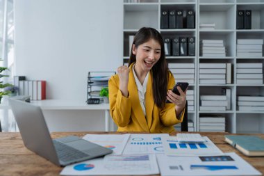 Modern ofis masasındaki akıllı telefonu iş raporlarıyla kontrol ederken başarıyı kutlayan heyecanlı genç iş kadını.