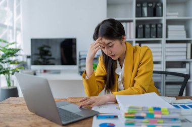 Sarı ceketli stresli iş kadını, modern ofis ortamında kağıtlar ve çizelgelerle dolu dizüstü bilgisayarda çalışıyor..