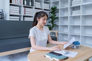 Finansal belgeleri analiz eden ve modern oturma odasındaki kahve masasına ev yatırımı planlayan genç bir kadın..