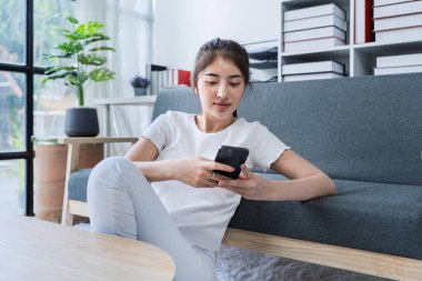 Modern oturma odasındaki akıllı telefondan keyif alan genç bir kadın rahat dekorlu ve doğal ışıklandırmalı..