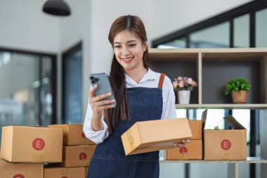 Çevrimiçi iş yapan genç bir kadın akıllı telefon kullanıyor, kargo için paket hazırlıyor ve müşteri adreslerini kutudan alıyor..
