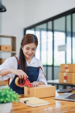 Genç bir kadın, kargo malzemeleriyle çevrili modern bir ofis çalışma yerinde bant dolu kutuları mutlu bir şekilde paketliyor..
