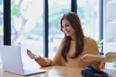 Gülümseyen genç bir kadın, neşeli bir e-öğrenme videosuyla meşgul. Kulaklıklı bir kitabı yakındaki tahta bir masada tutarken el kol hareketi yapıyor..