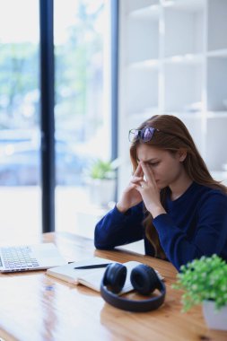 Masasında dizüstü bilgisayar, not defteri, kulaklık ve elleri hayal kırıklığı içinde yüzüne yapışan e-öğrenim çalışmalarından bunalmış genç bir kadın..