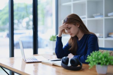 Masasında dizüstü bilgisayar, not defteri, kulaklık ve elleri hayal kırıklığı içinde yüzüne yapışan e-öğrenim çalışmalarından bunalmış genç bir kadın..