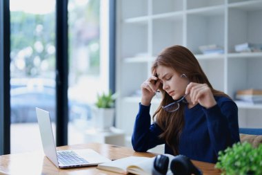 Masasında dizüstü bilgisayar, not defteri, kulaklık ve elleri hayal kırıklığı içinde yüzüne yapışan e-öğrenim çalışmalarından bunalmış genç bir kadın..