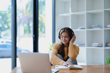 Genç bir kadın masasında dinlendirici bir müzik molası verirken kulaklık kullanıyor, deftere yazıyor ve sakin bir ilham için dizüstü bilgisayarı üzerinde çalışıyor..