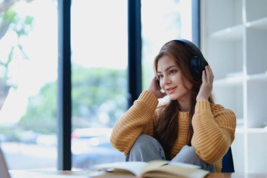 Genç bir kadın masasında dinlendirici bir müzik molası verirken kulaklık kullanıyor, deftere yazıyor ve sakin bir ilham için dizüstü bilgisayarı üzerinde çalışıyor..