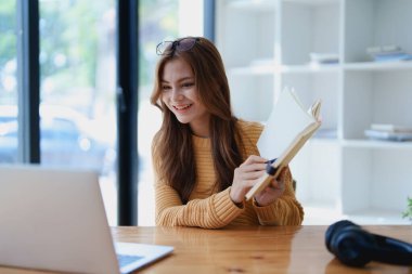 Gülümseyen genç bir kadın, neşeli bir e-öğrenme videosuyla meşgul. Kulaklıklı bir kitabı yakındaki tahta bir masada tutarken el kol hareketi yapıyor..